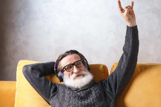 Man with white beard, wearing headsets, sitting on couch and making a Shaka sign with his left hand. 