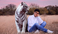 young man sitting in a field with a big tiger sitting next to him